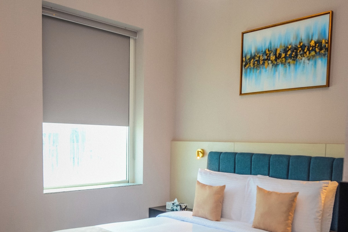 Ein ordentlich bezogenes Bett mit blauem Kopfteil, einem Nachttisch und einem Bild an der Wand.