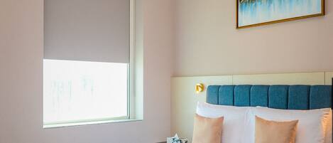 A hotel room with a bed, a window, a painting, and a bedside table with a water bottle and a box.