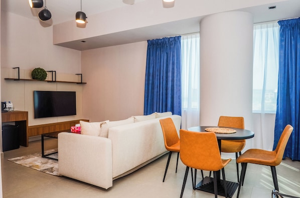 A modern living room with a sofa, dining table, and chairs, a flat-screen TV, and blue curtains. - VOGO Grand Hotel formerly Best Western Plus Academic City (Dubai)