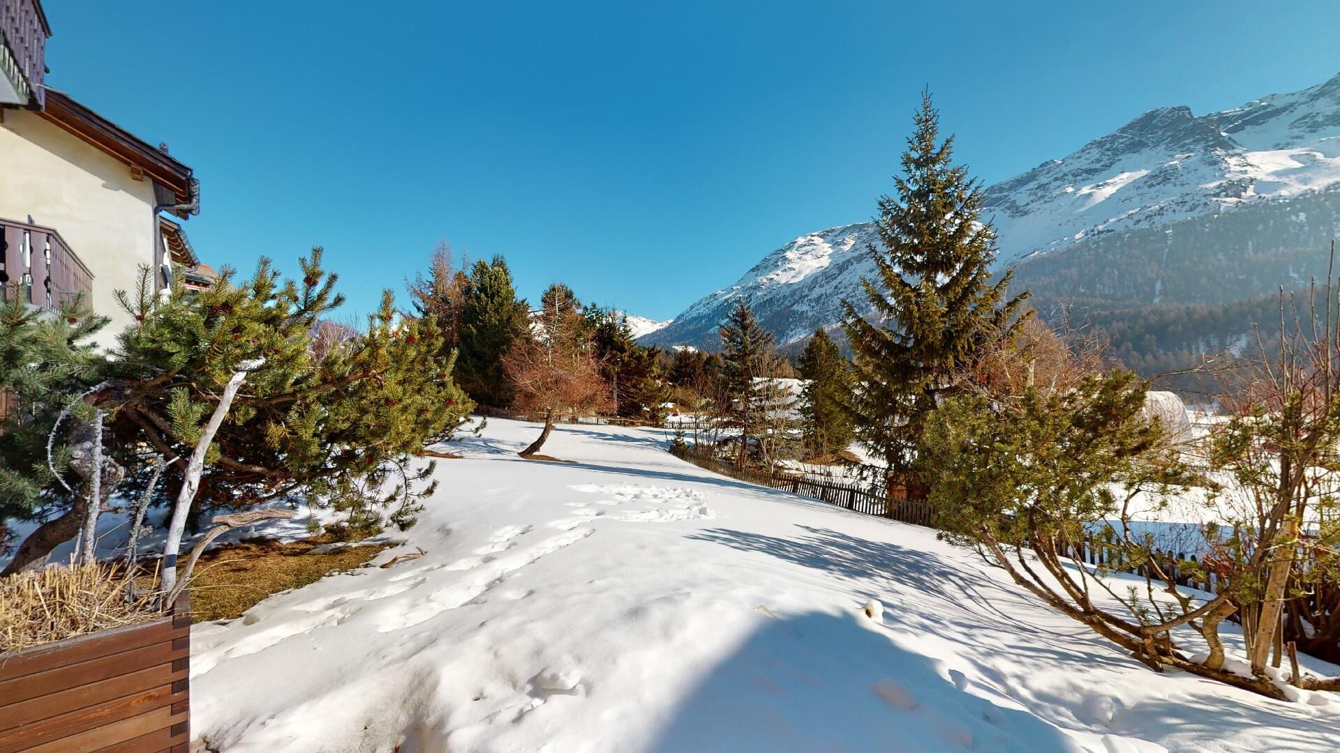 A snow-covered path leading to a building with a wooden deck, surrounded by evergreen trees and a mountain backdrop.