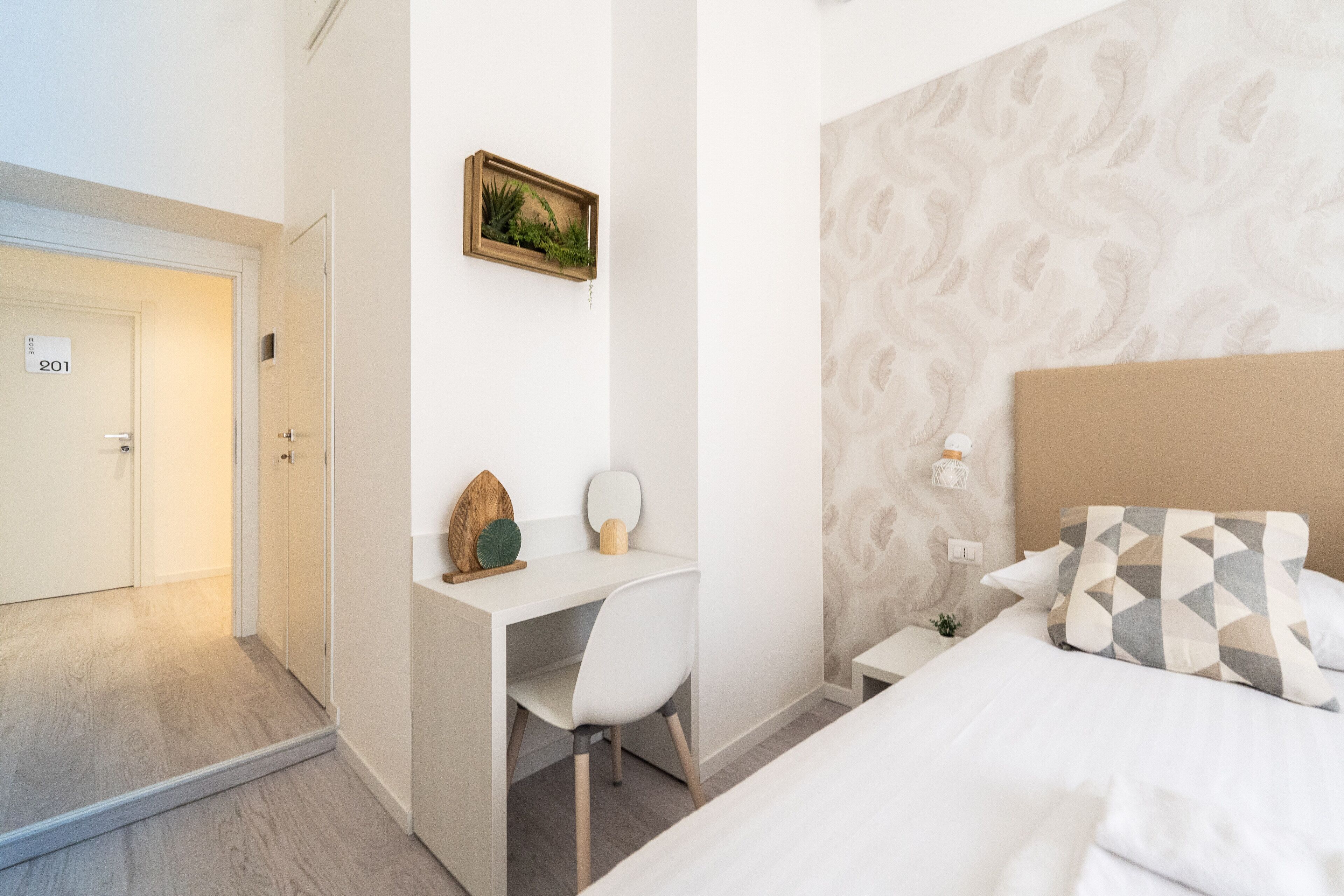 A hotel room with a bed, a desk with a chair, a small table, and a wall-mounted shelf with decorative items.