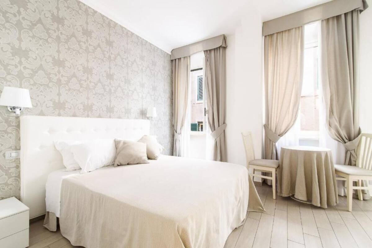 A hotel room with a large bed, a bedside table with a lamp, a chair, a small table, and a window with curtains.