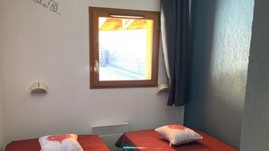 Deux lits simples avec une literie rouge et des oreillers blancs, une fenêtre avec des rideaux, un luminaire fixé au mur et un radiateur.