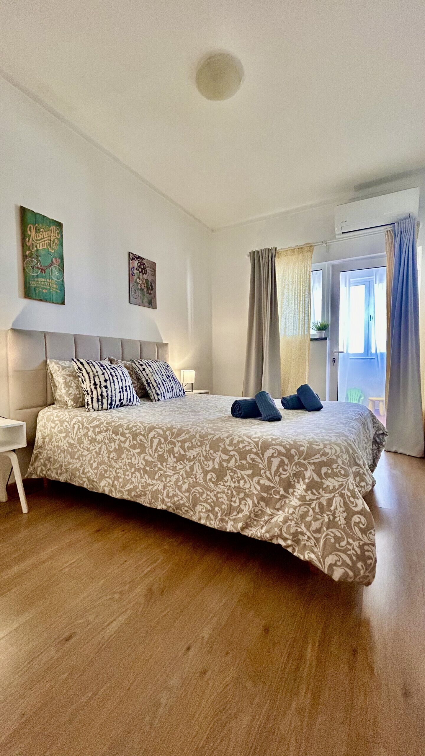 A bedroom with a bed, a nightstand, a wall-mounted air conditioner, and a window with curtains.