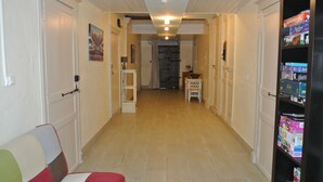 Un pasillo con piso de baldosas, paredes blancas e iluminación empotrada en el techo. A la izquierda, una puerta; a la derecha, una pequeña mesa con lámpara y una repisa con objetos.