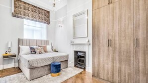 A bedroom with a bed, bedside tables, a dresser, a mirror, a fireplace, and a wardrobe. - Stylish Chelsea Apartment on King’s Road (London)