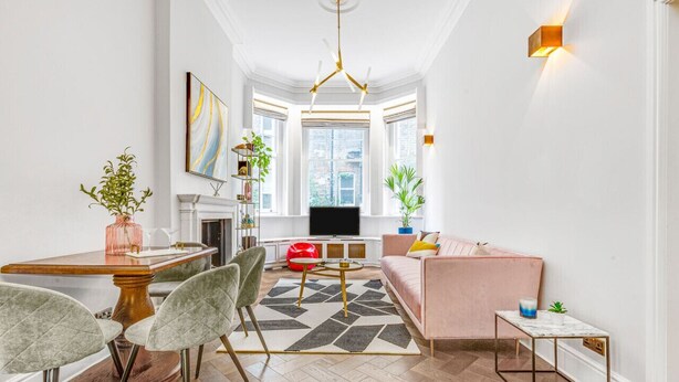 A modern living room with a sofa, a coffee table, and a dining area with chairs and a table. There is a TV mounted on the wall and a fireplace. - Stylish Chelsea Apartment on King’s Road (London)