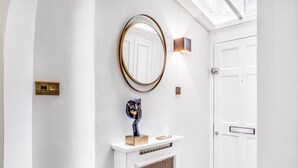 A hallway with a white door, a round mirror with a gold frame, a wall-mounted light, and a small sculpture on a white console table. - Stylish Chelsea Apartment on King’s Road (London)