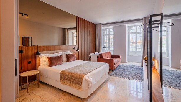 Ein modernes Hotelzimmer mit einem großen Bett, einem Schreibtisch, einem Sessel und Blick auf die Stadt durch große Fenster.