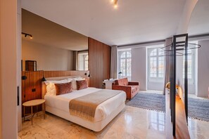 Ein modernes Hotelzimmer mit einem großen Bett, einem Schreibtisch, einem Sessel und Blick auf die Stadt durch große Fenster.