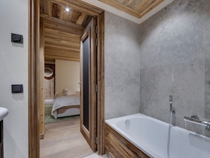 A modern bathroom with a bathtub, a shower, and a wooden door leading to a bedroom.