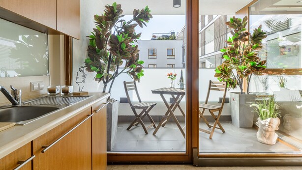 Une cuisine moderne avec des meubles en bois, un évier en acier inoxydable et un coin repas comprenant une table et des chaises. On y trouve également des plantes en pot et, grâce à la porte vitrée, on peut admirer la vue sur l’extérieur.