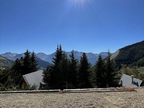 Paisagem montanhosa sob um céu claro. Árvores e um edifício com telhado inclinado ao primeiro plano.