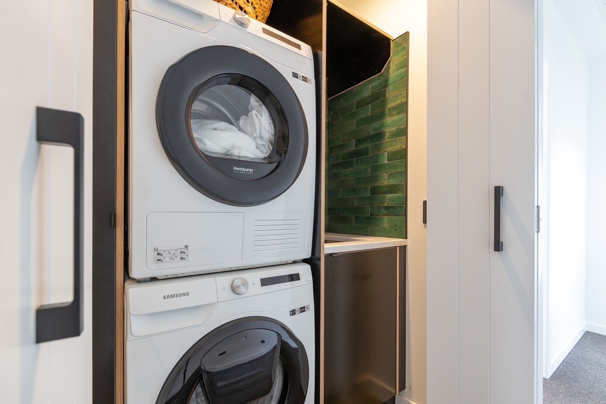 A compact laundry area with a front-loading washer and dryer, a basket on top, and a green brick backsplash.