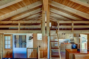 L’intérieur d’un chalet rustique, avec un plafond haut, des poutres en bois, un coin cuisine et un escalier menant à une mezzanine.