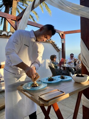 Un chef préparant des plats à l’extérieur, avec une vue sur l’espace réservé aux repas et les convives.