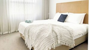 Ein ordentlich bezogenes Bett mit weißen Leinen, einem beige gefärbten Kopfteil und einer weißen Tagesdecke.