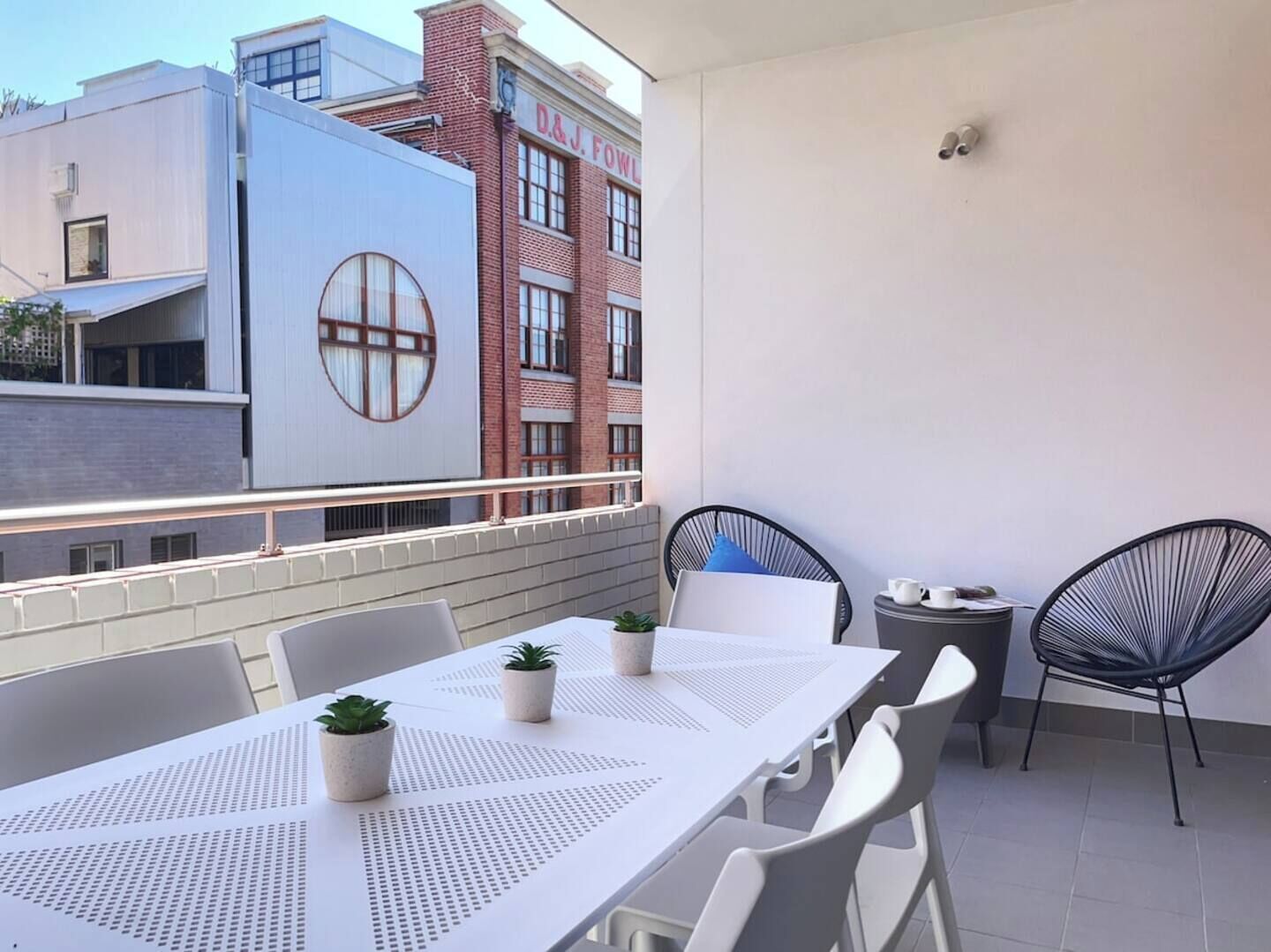 A balcony with a white table and chairs, potted plants, and a view of buildings with a red brick facade and a white building with a circular window.