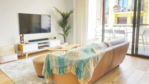 Ein modernes Wohnzimmer mit Sofa, Fernseher, einer Pflanze und einem Balkon mit Sitzgelegenheit.