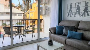 En balkon med et bord og stole, en grå sofa med blå puder og en glaskrukke med stueplanter.