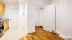 A modern kitchen with wooden cabinets, a washing machine, and a hallway with wooden flooring leading to a bedroom. - 2 Bed Apartment Right on Trafalgar Square  (London)