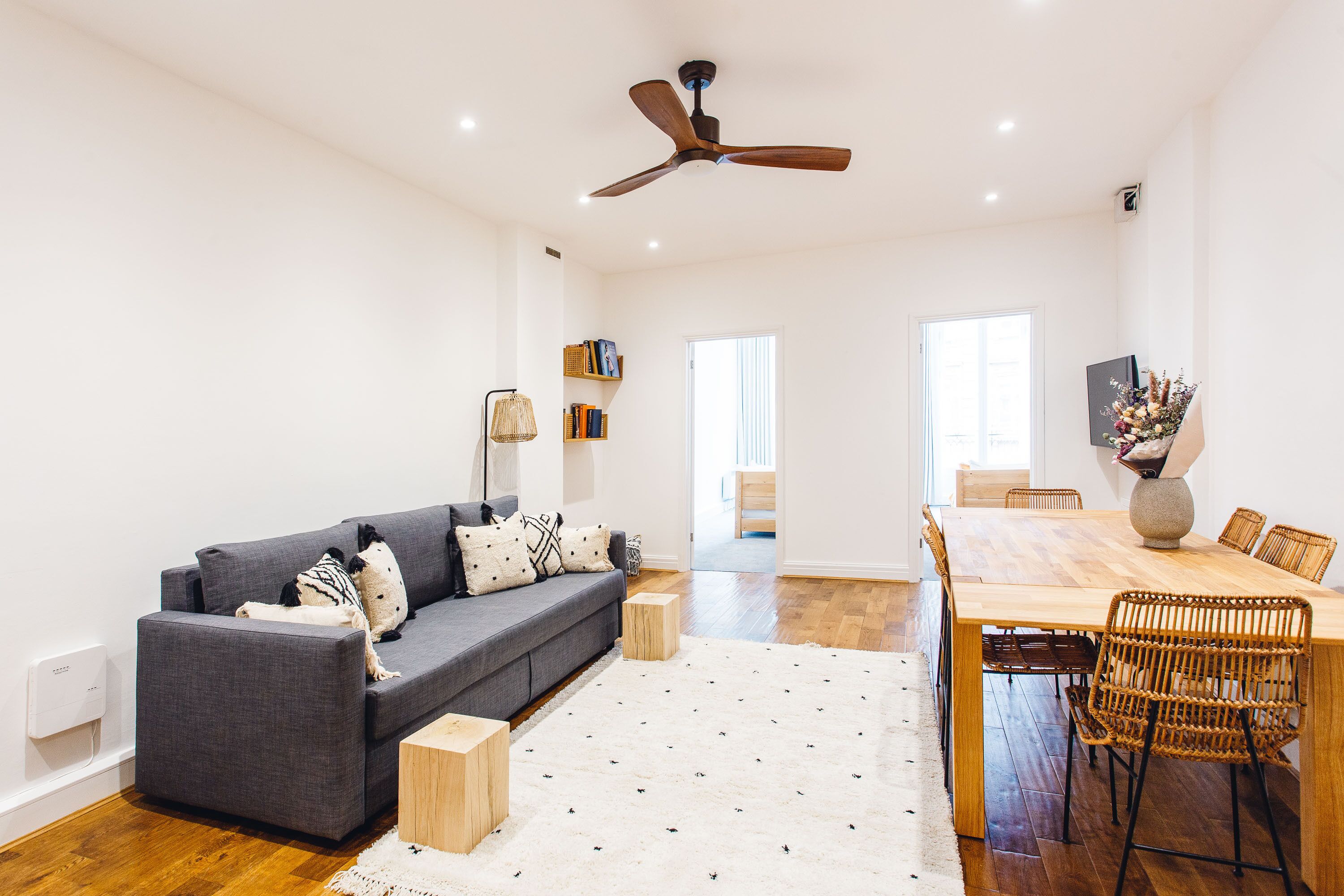 A modern living room with a grey sofa, wooden coffee table, and a dining area with a wooden table and wicker chairs.