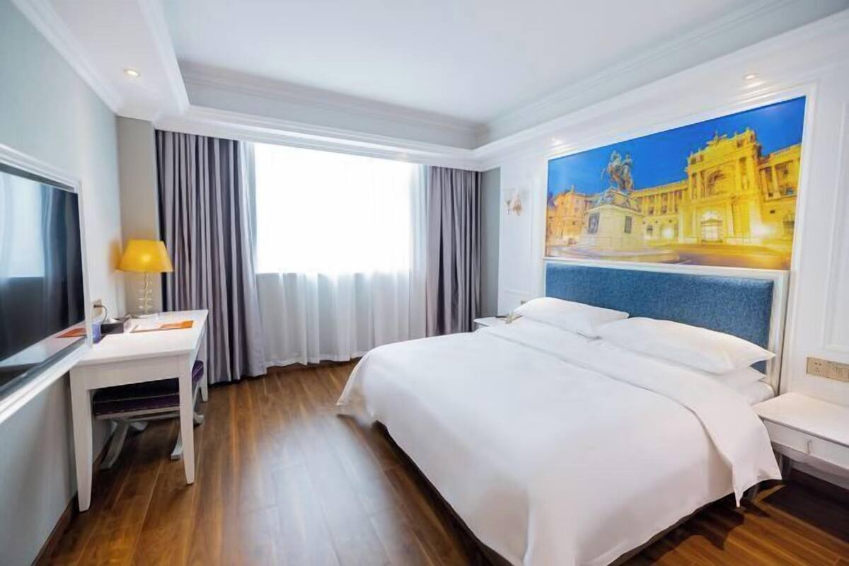 A hotel room with a bed, a desk, a chair, a TV, and a large window with curtains.