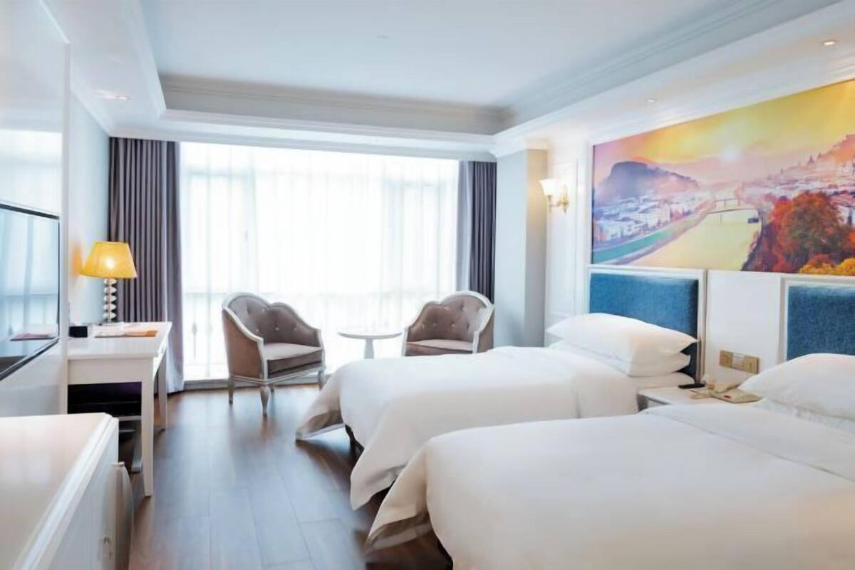 A hotel room with two beds, a desk, a chair, and a large window with curtains.