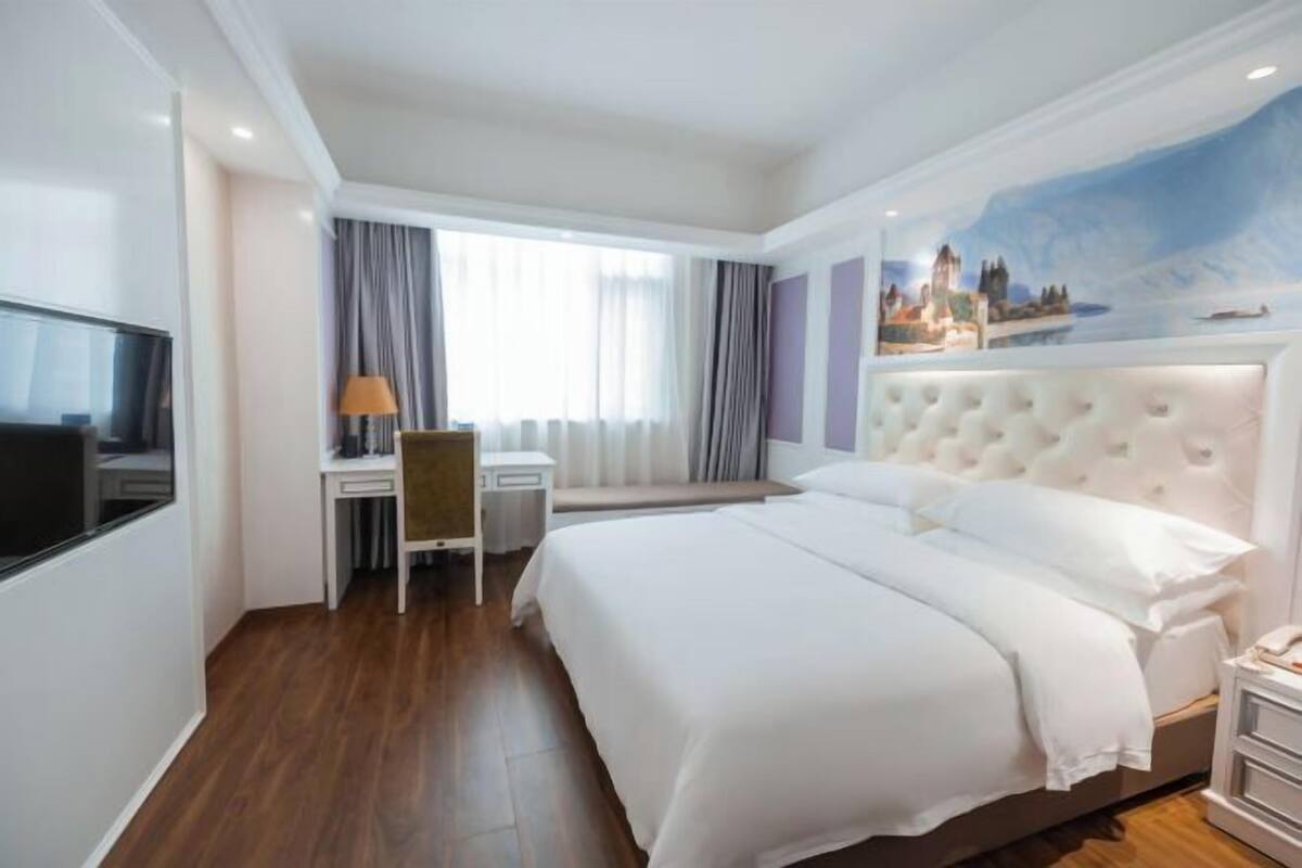 A hotel room with a bed, a desk, a chair, a TV, and a painting on the wall.
