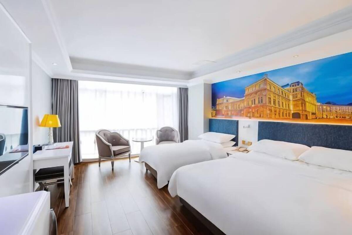 A hotel room with two beds, a desk, a chair, a TV, and a painting of a building on the wall.