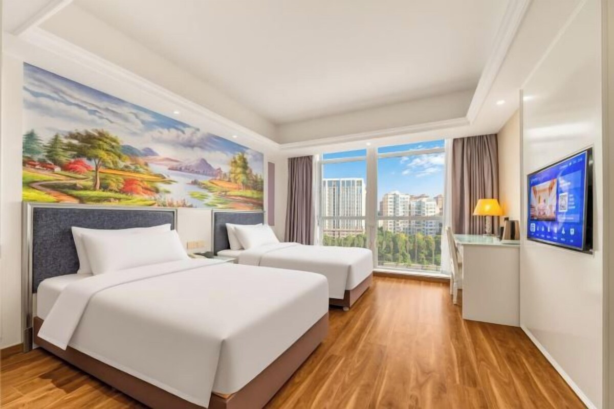 A hotel room with two beds, a large window with a city view, a flat-screen TV, and a painting on the wall.
