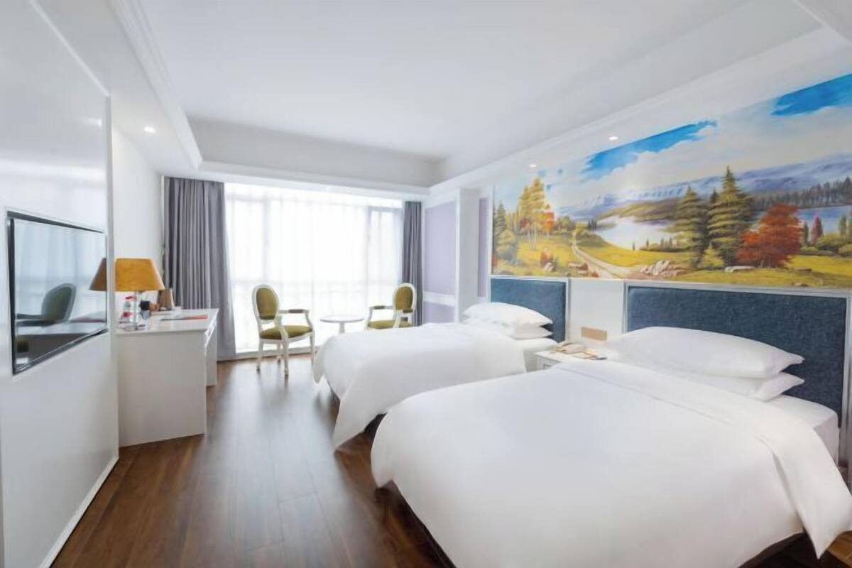 A hotel room with two beds, a desk with a lamp, a chair, and a large painting on the wall.