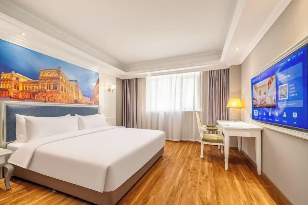 A hotel room with a large bed, a desk, a chair, a TV, and a mural of a building.