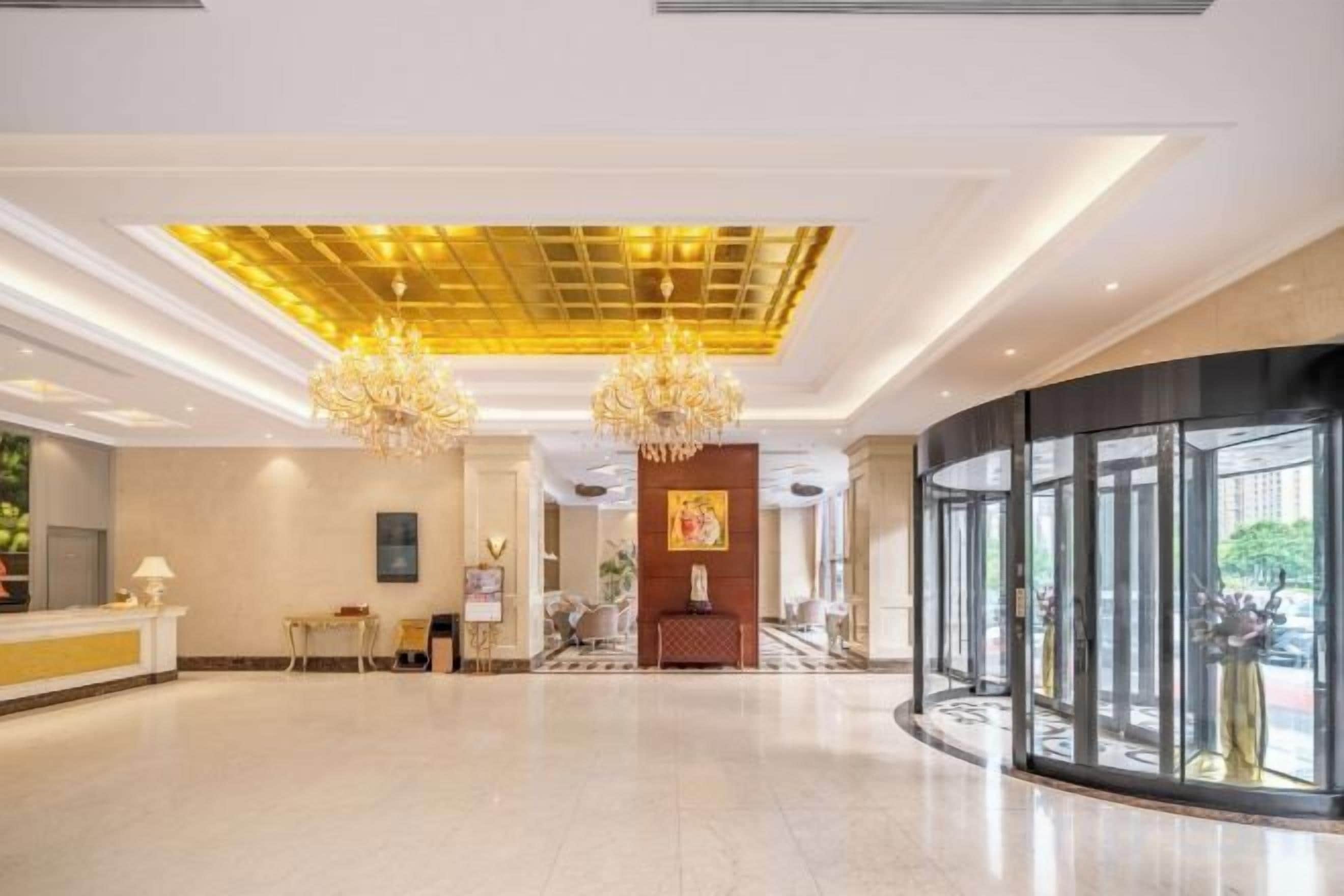 A spacious lobby with a reception desk, chandeliers, and a large glass wall.