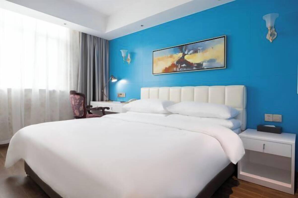 A modern hotel room with a large bed, a nightstand, a desk, and a painting on the wall.