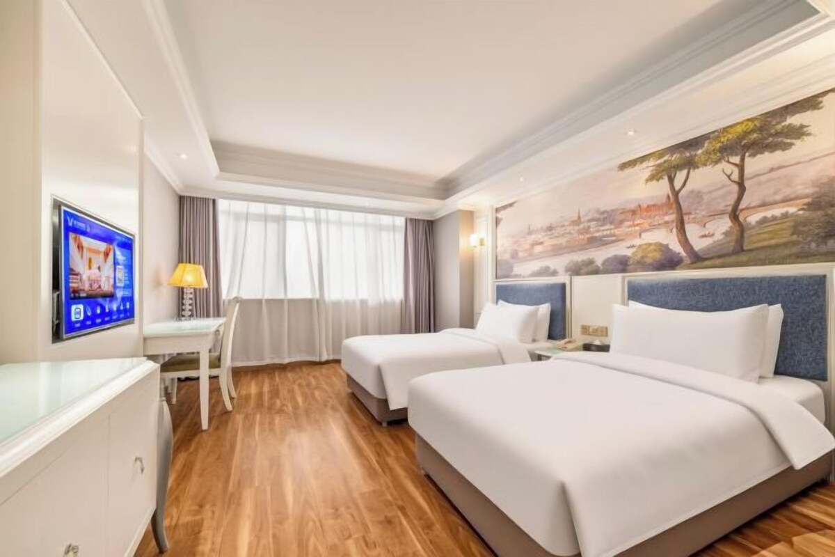 A hotel room with two beds, a desk, a TV, and a painting on the wall.