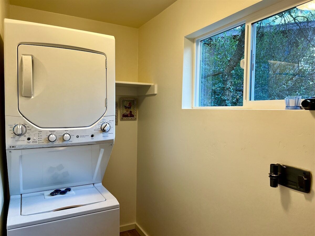 A compact laundry room with a front-loading washer and dryer, a small shelf above the dryer, and a window with a view of greenery outside.