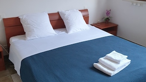 A neatly made bed with white linens and a blue blanket, a wooden nightstand with a small potted plant, and a wall-mounted light fixture.