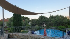 A wooden deck with a stone wall, a metal railing, and a view of a swimming pool and trees.