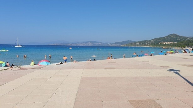 A beach with a tiled promenade, clear blue sea, and distant hills.