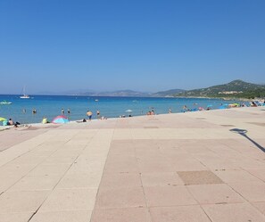 A beach with a tiled promenade, clear blue sea, and distant hills.