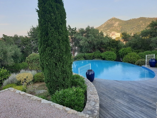 A swimming pool with a wooden deck, surrounded by greenery and a mountainous backdrop.