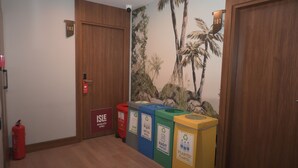 Un couloir avec des portes en bois, un extincteur, des poubelles de recyclage et une fresque représentant des palmiers.