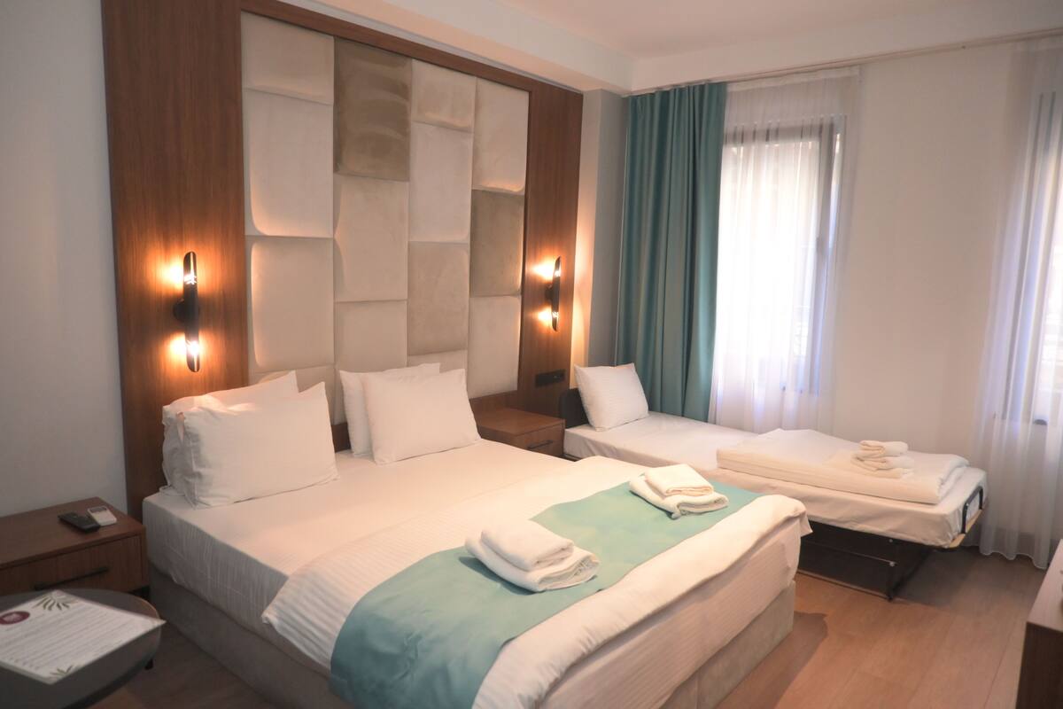 A hotel room with two beds, a wooden headboard, a nightstand, a desk, and a window with curtains.