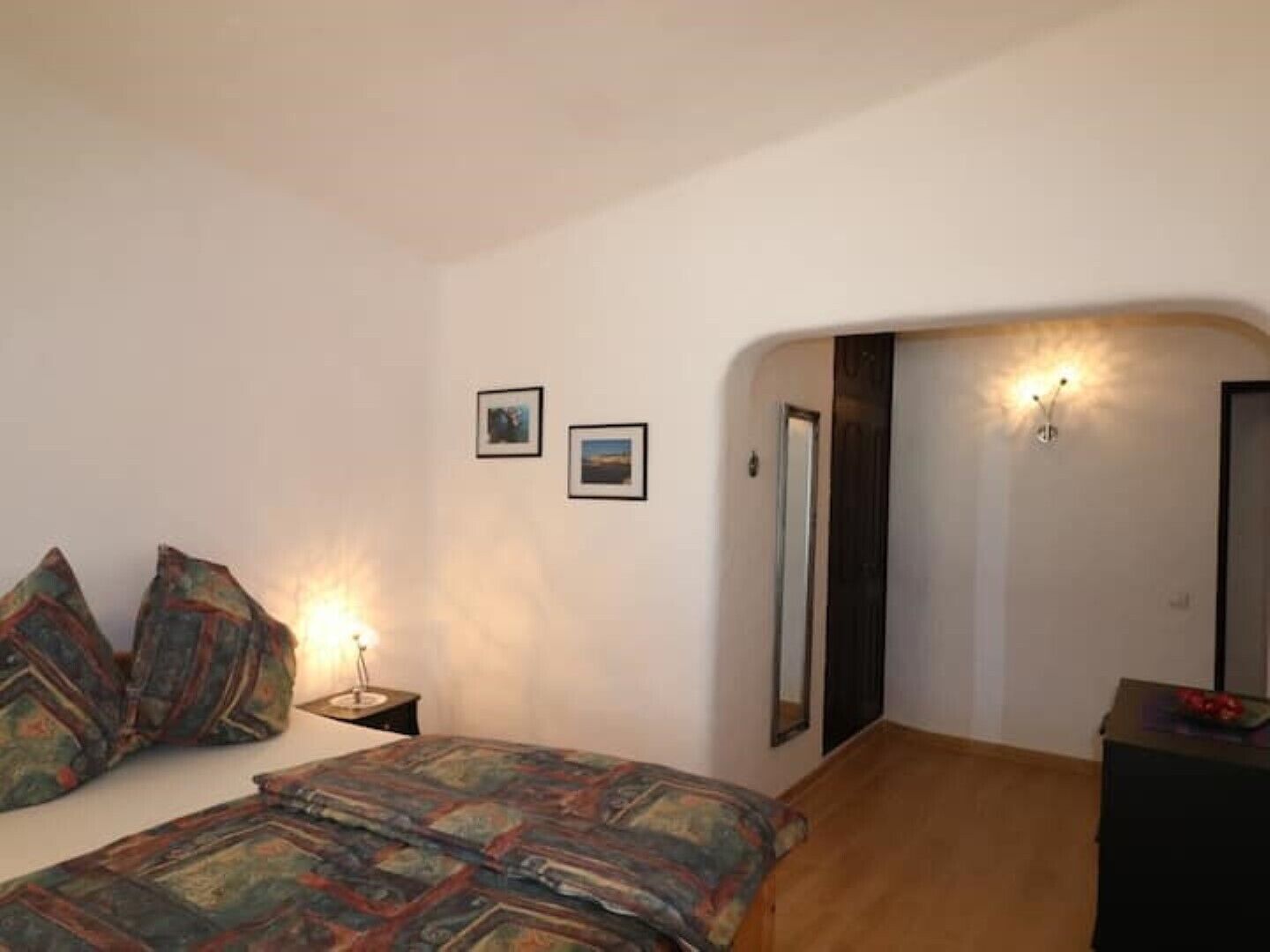 A bedroom with a bed, two pillows, a nightstand, a wall-mounted lamp, and two framed pictures.