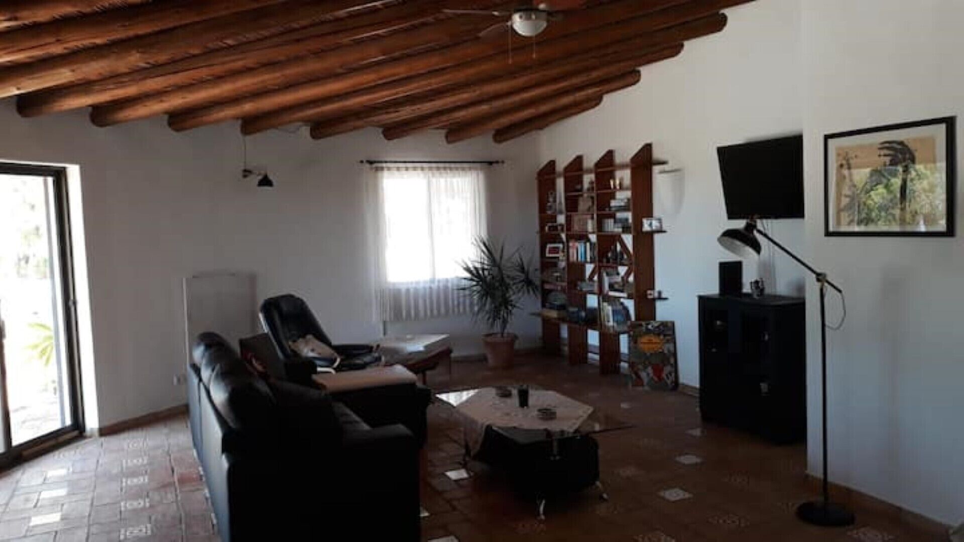 A living room with a wooden ceiling, a sofa, a coffee table, a bookshelf, a TV, and a window with curtains.