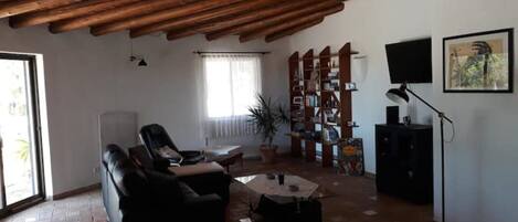 A living room with a wooden ceiling, a sofa, a coffee table, a bookshelf, a TV, and a window with curtains.