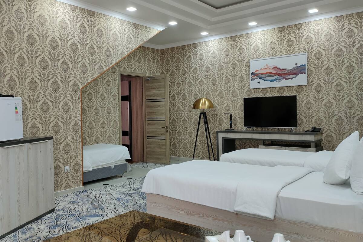 A hotel room with a patterned wallpaper, a bed, a desk with a TV, and a refrigerator.