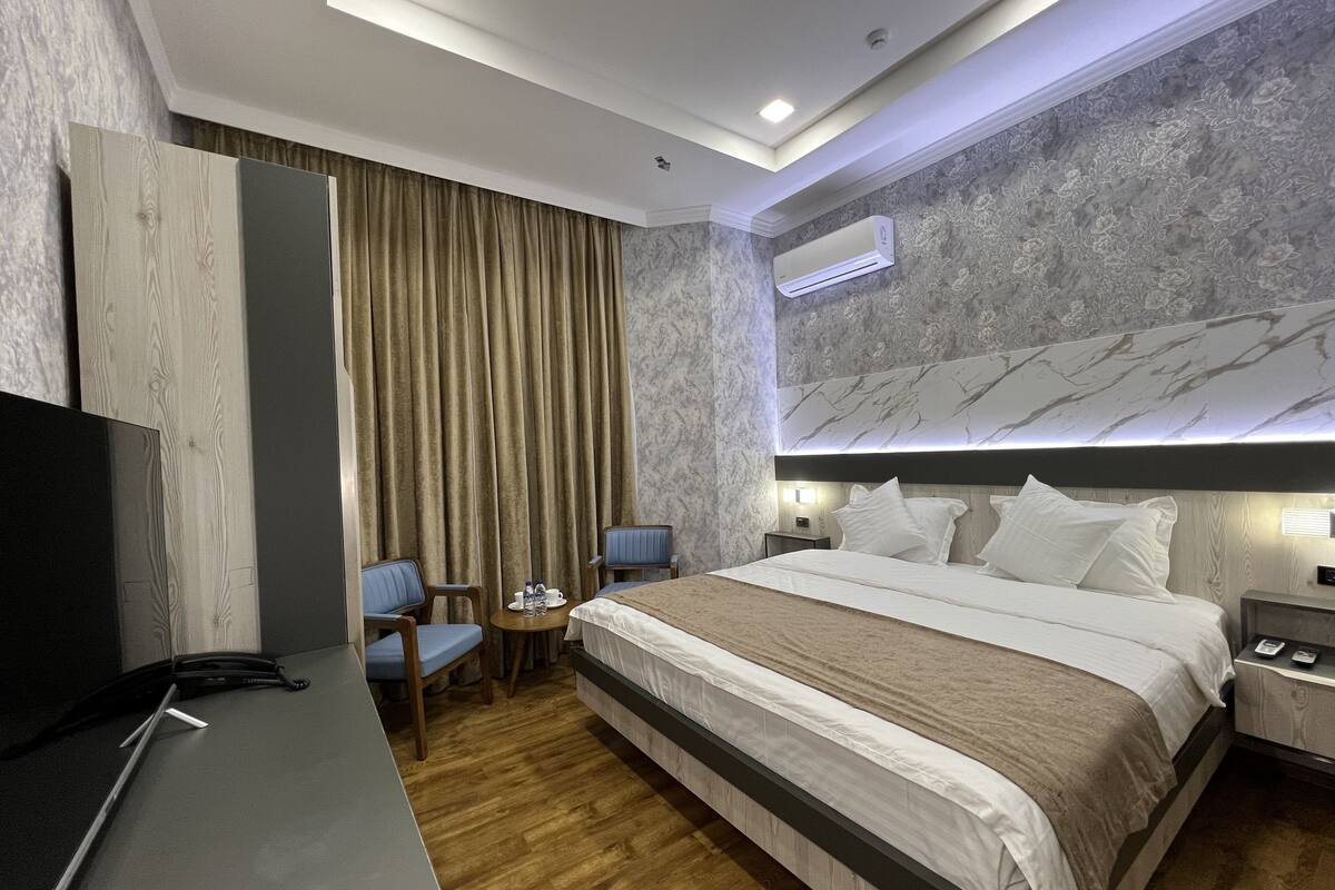 A modern hotel room with a large bed, a desk, two chairs, an air conditioning unit, and a television.