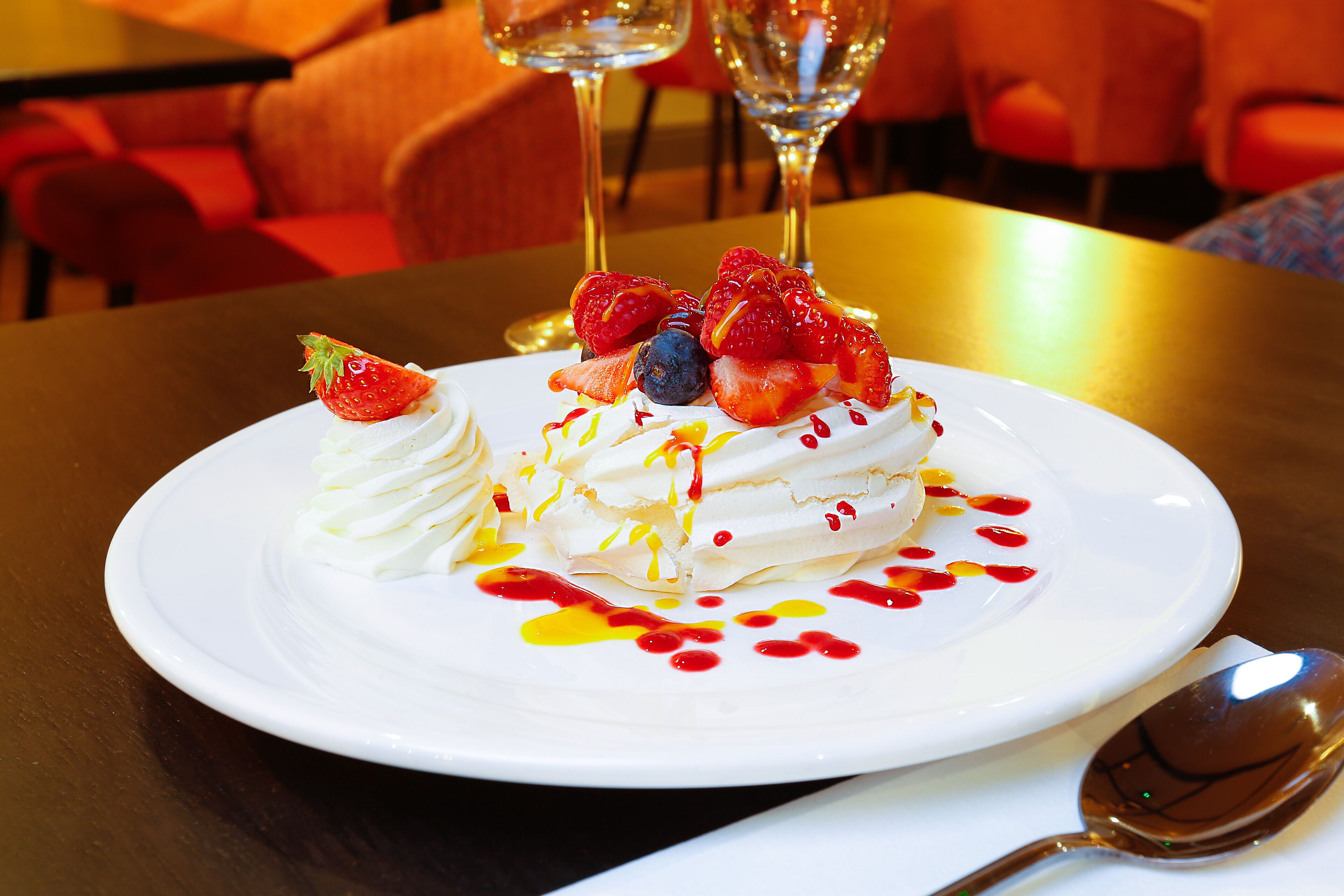 A dessert plate with whipped cream, strawberries, and blueberries, accompanied by a spoon and two glasses of white wine on a table.